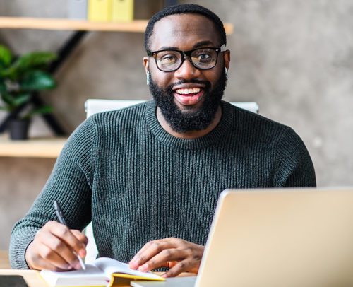 A Black man sitting in front of his laptop, writing but looking up and smiling at the camera