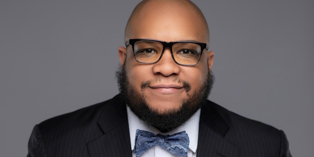 Photo shows a man with glasses and a beard in a black suit with a gray bowtie. 